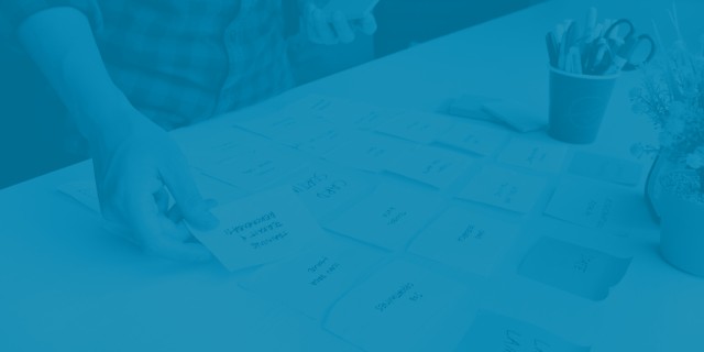 Person arranging handwritten sticky notes across a table during a collaborative planning or UX workshop, with office supplies and plants in the background. The image is covered by a semi-transparent blue overlay, making the scene appear muted and monochromatic.
